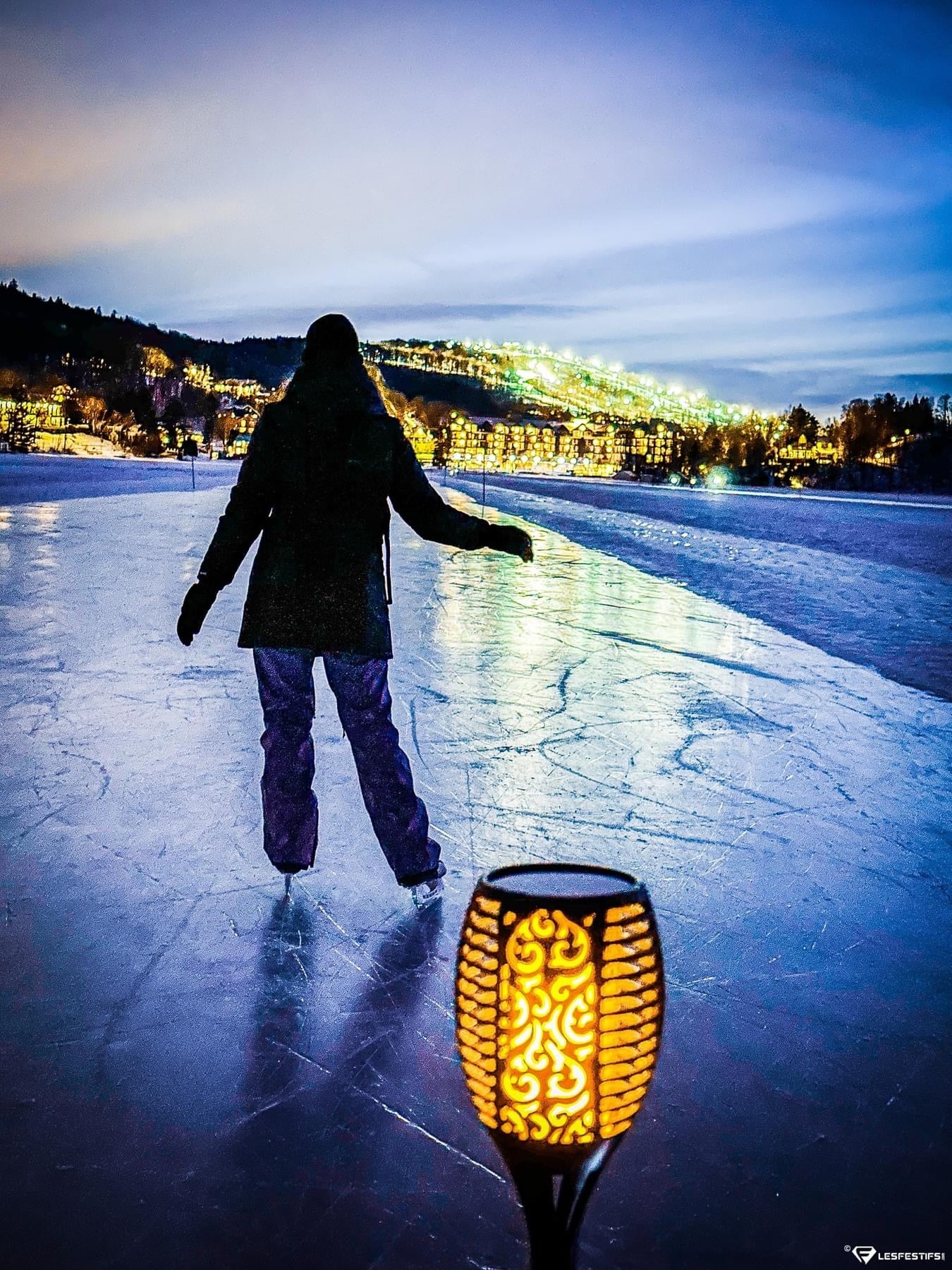 A person in a dark winter coat and pants skates on a frozen lake at dusk, with a glowing decorative BigM 96 LED Flickering Flame Solar Tiki Torch Lights in the foreground and city lights on hills in the background.