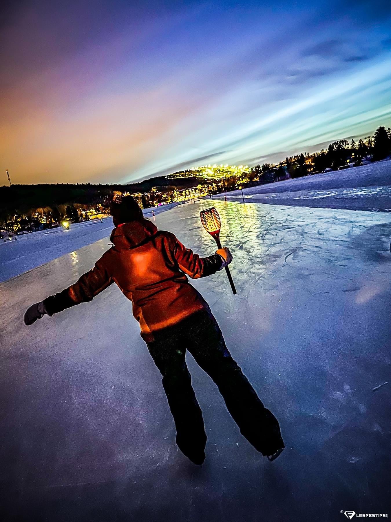 A person in a red-orange hooded jacket and dark pants stands on a frozen lake at dusk at municipalité de lac-beauport, Quebec, holding a glowing BigM 96 LED Flickering Flame Solar Tiki Torch Lights. The background shows a snowy landscape, distant lights, and a blue-purple sky.