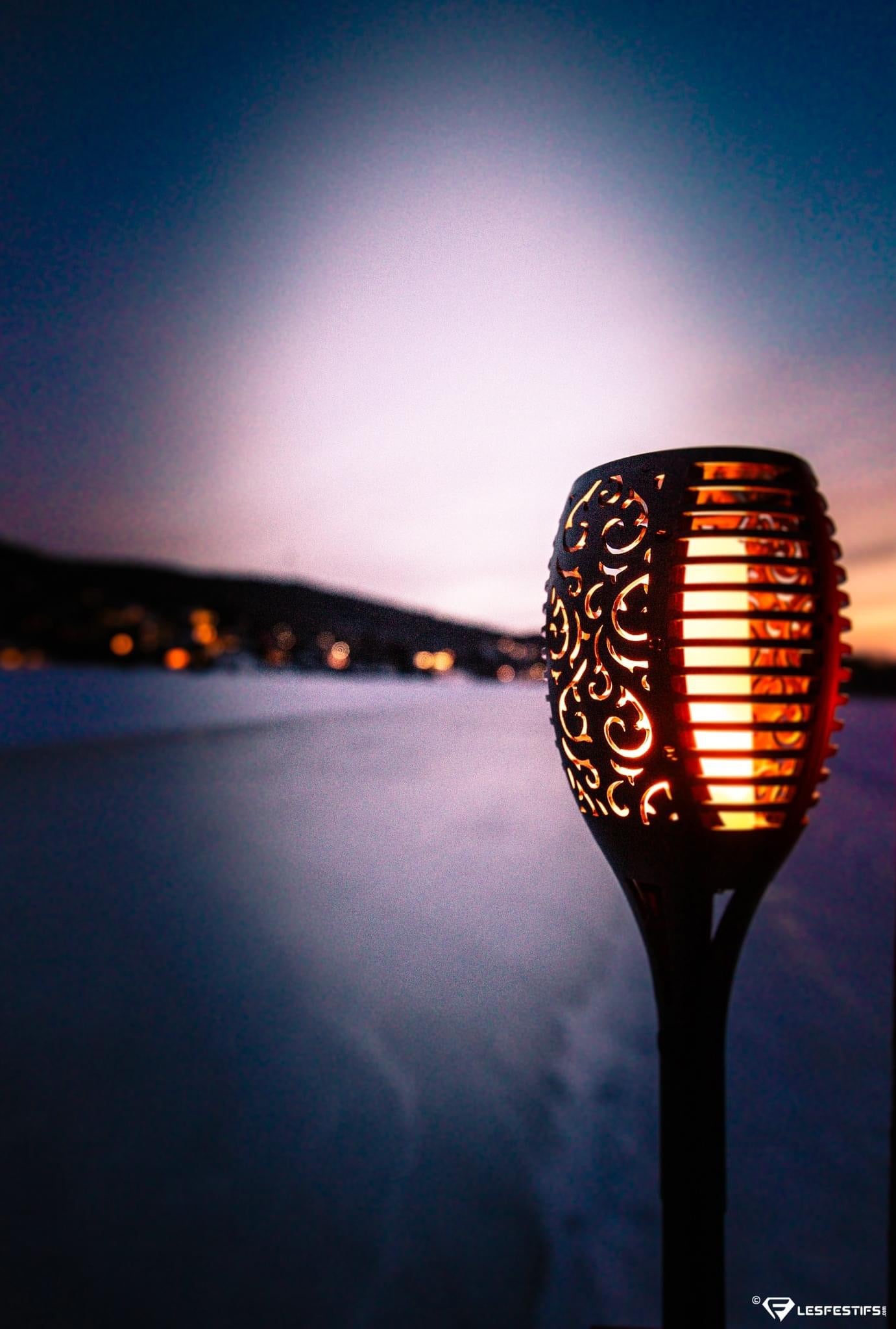 A decorative outdoor BigM 96 LED Flickering Flame Solar Tiki Torch Lights with intricate cutout scrollwork and glowing warm orange-red light, photographed against a blurred twilight beach or lake background with distant lights.