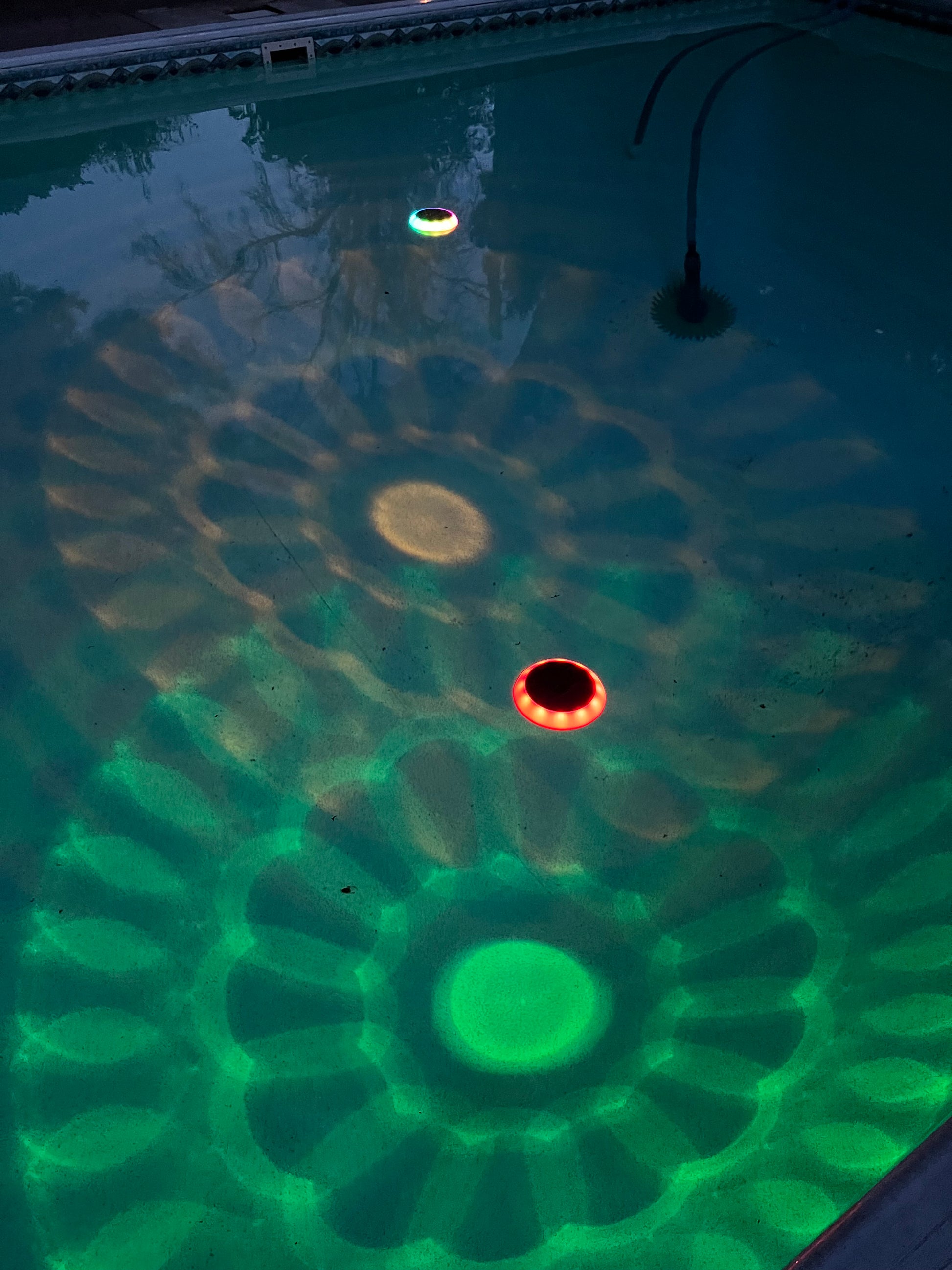 BigM Solar LED Floating Pool Light with RGB Colour Changing feature shining on a pool. A close-up view of a swimming pool waterline showing a green and blue LED light pattern projected onto the water surface, with a circular opening in the center.