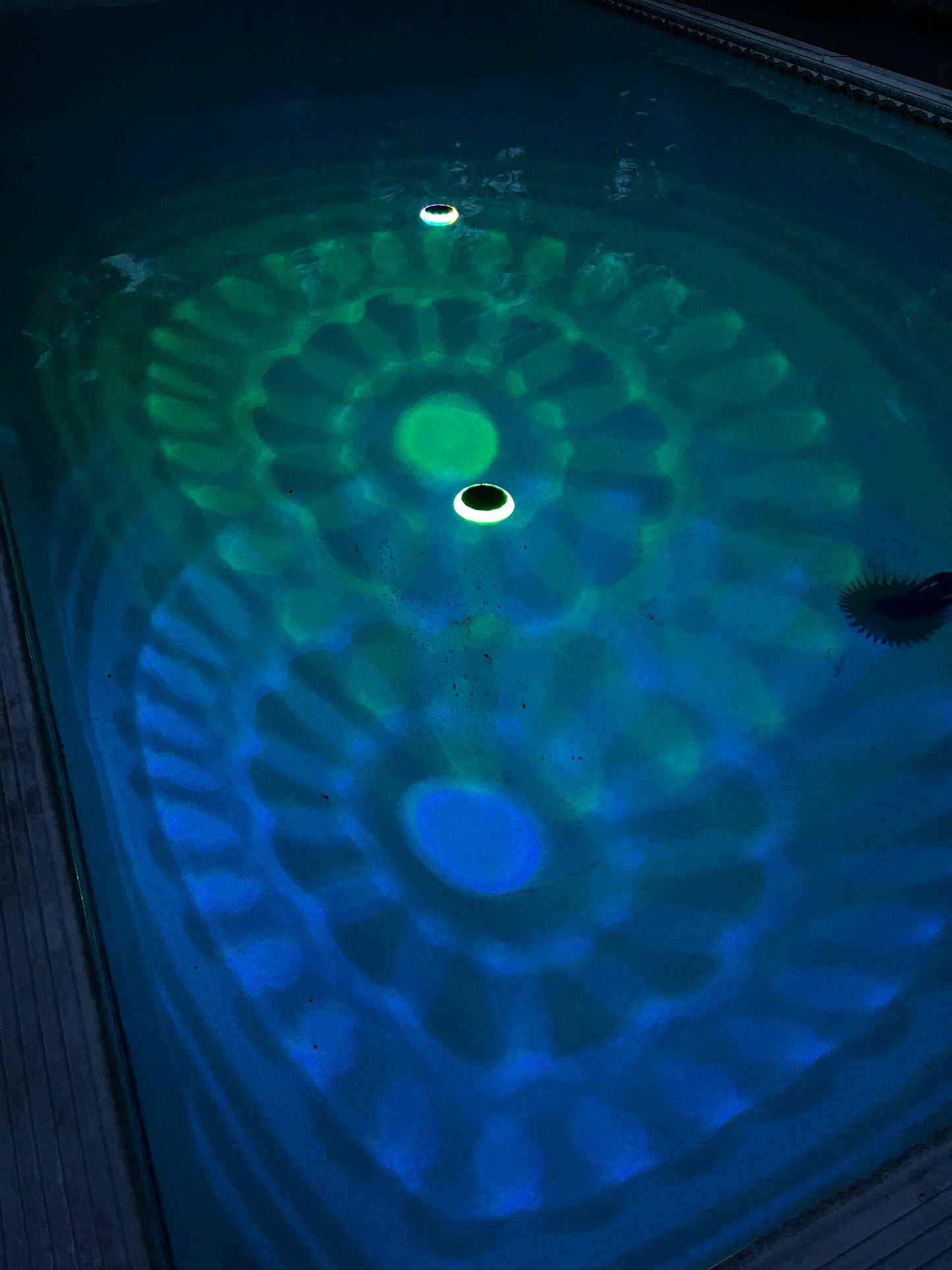 BigM Solar LED Floating Pool Light with RGB Colour Changing feature shining on a pool. A close-up view of a swimming pool waterline showing a green and blue LED light pattern projected onto the water surface, with a circular opening in the center.