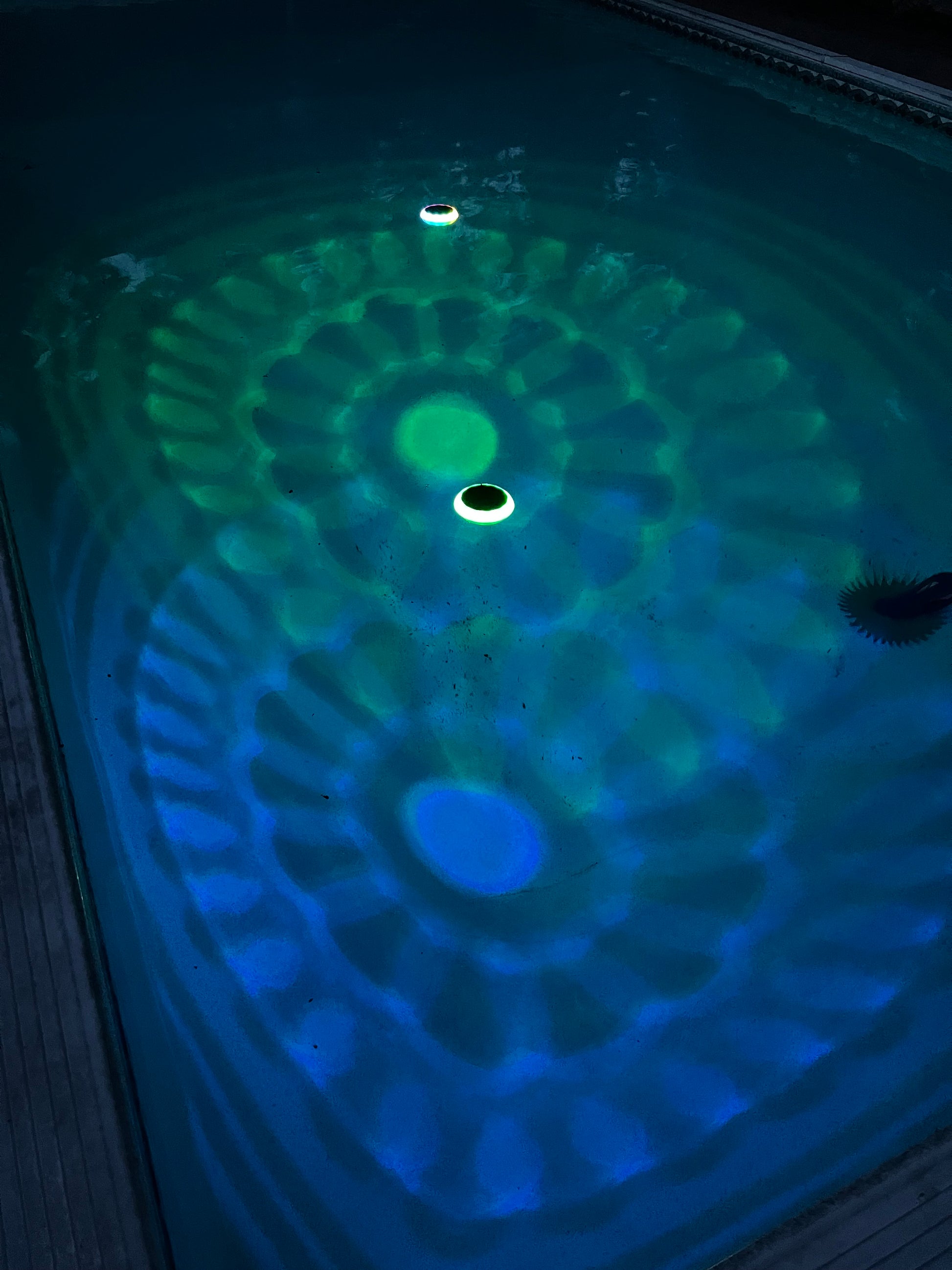 BigM Solar LED Floating Pool Light with RGB Colour Changing feature shining on a pool. A close-up view of a swimming pool waterline showing a green and blue LED light pattern projected onto the water surface, with a circular opening in the center.