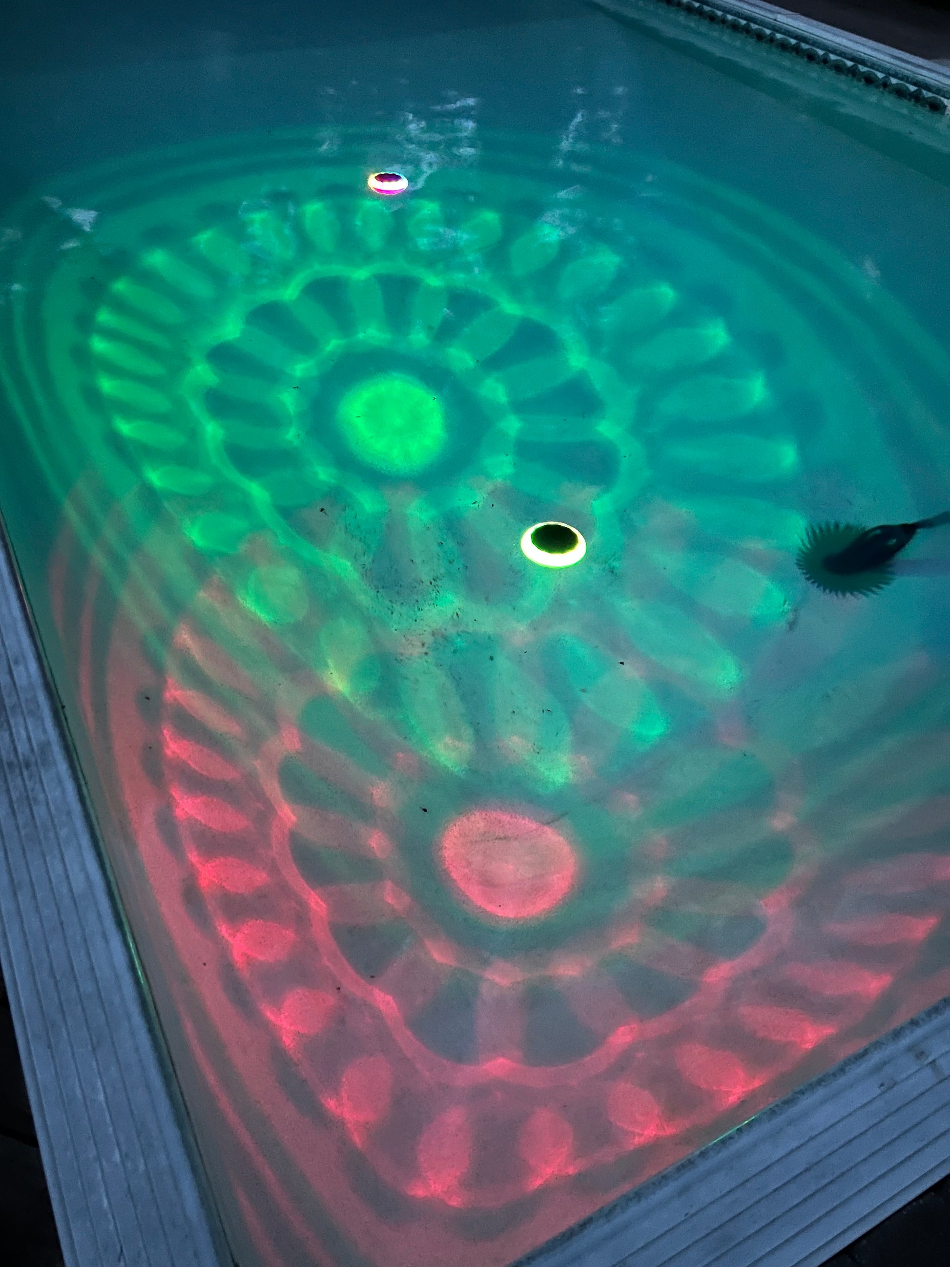 BigM Solar LED Floating Pool Light with RGB Colour Changing feature shining on a pool. A close-up view of a swimming pool waterline showing a green and red LED light pattern projected onto the water surface, with a circular opening in the center.