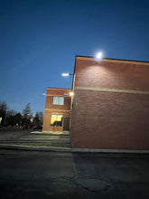 Load image into Gallery viewer, Image of BigM 700w solar parking lot lights lighting up a commercial building and pathways
